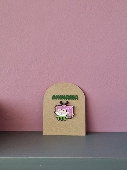 A white cute ghost popping out of a pink TV with green slime coming out of the bottom. Pin is mounted on a brown cardboard backing with the brand Animania written above it.