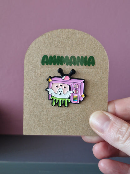 A white cute ghost popping out of a pink TV with green slime coming out of the bottom. Pin is mounted on a brown cardboard backing with the brand Animania written above it.