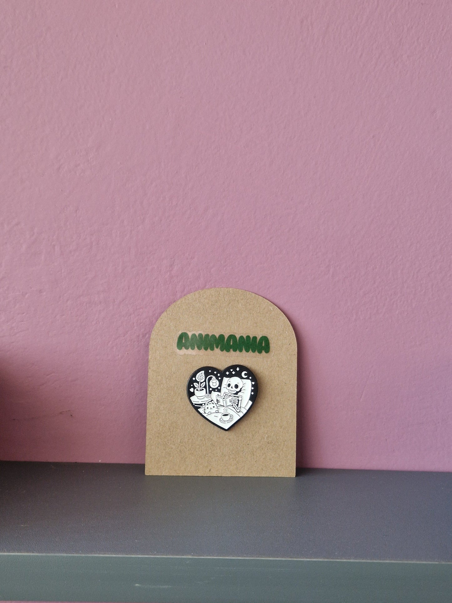 A black and white heart shaped pin of a skeleton reading a book in bed. Pin is mounted on a brown cardboard backing with the brand Animania written above it.