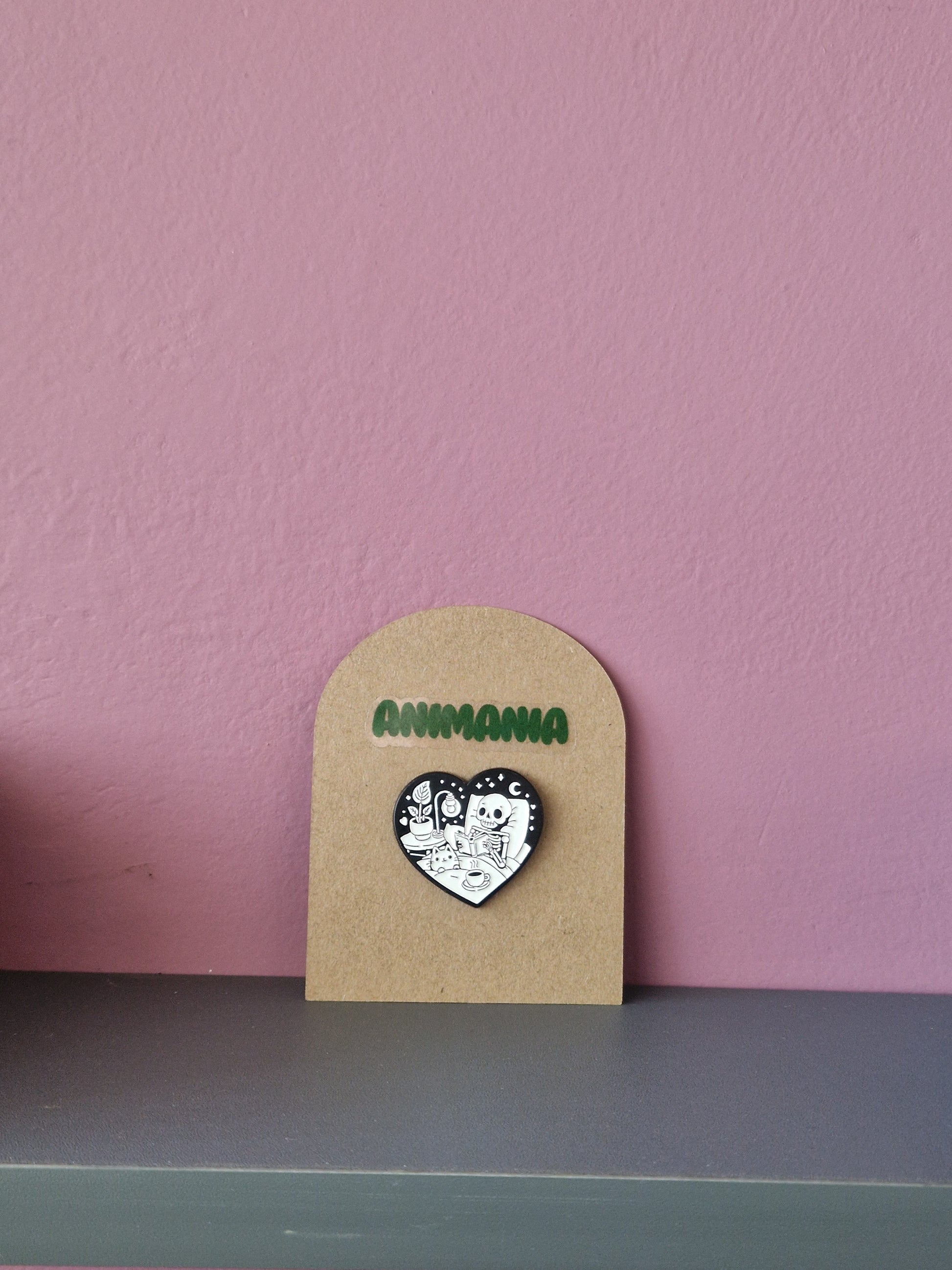A black and white heart shaped pin of a skeleton reading a book in bed. Pin is mounted on a brown cardboard backing with the brand Animania written above it.
