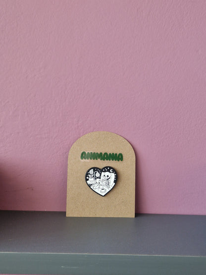 A black and white heart shaped pin of a skeleton reading a book in bed. Pin is mounted on a brown cardboard backing with the brand Animania written above it.
