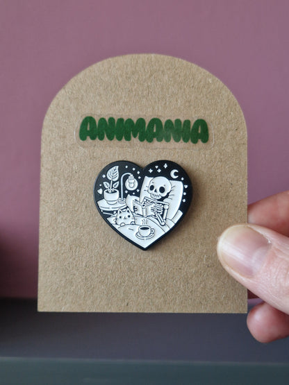 A black and white heart shaped pin of a skeleton reading a book in bed. Pin is mounted on a brown cardboard backing with the brand Animania written above it.