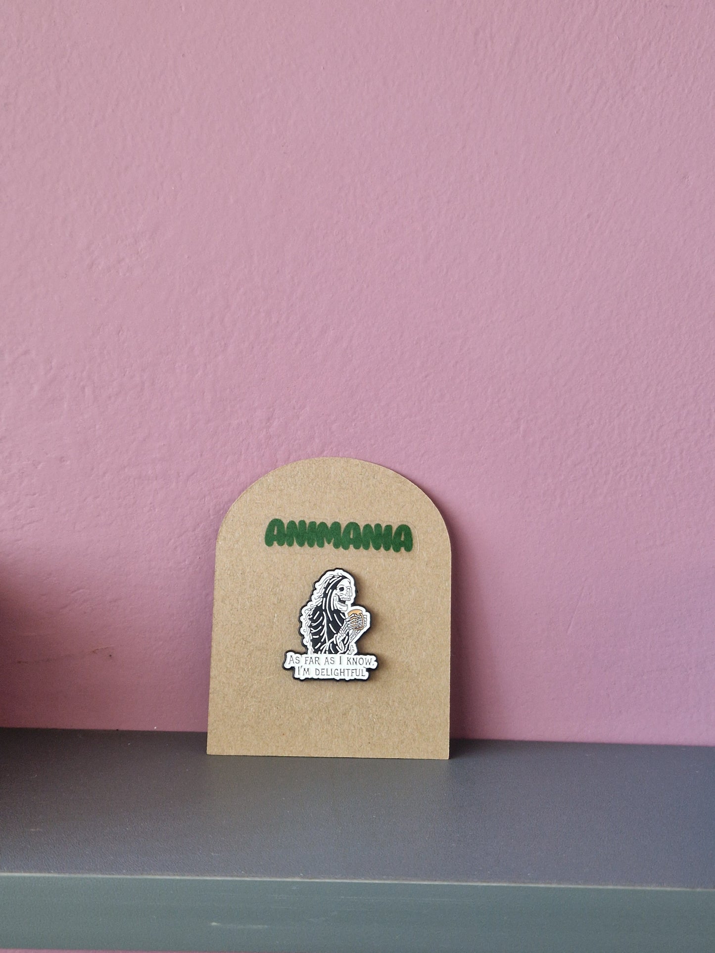 A black and white skeleton holding a cup of coffee with 'as far as i know, i'm delightful' written underneath it. Pin is mounted on a brown cardboard backing with the brand Animania written above it.
