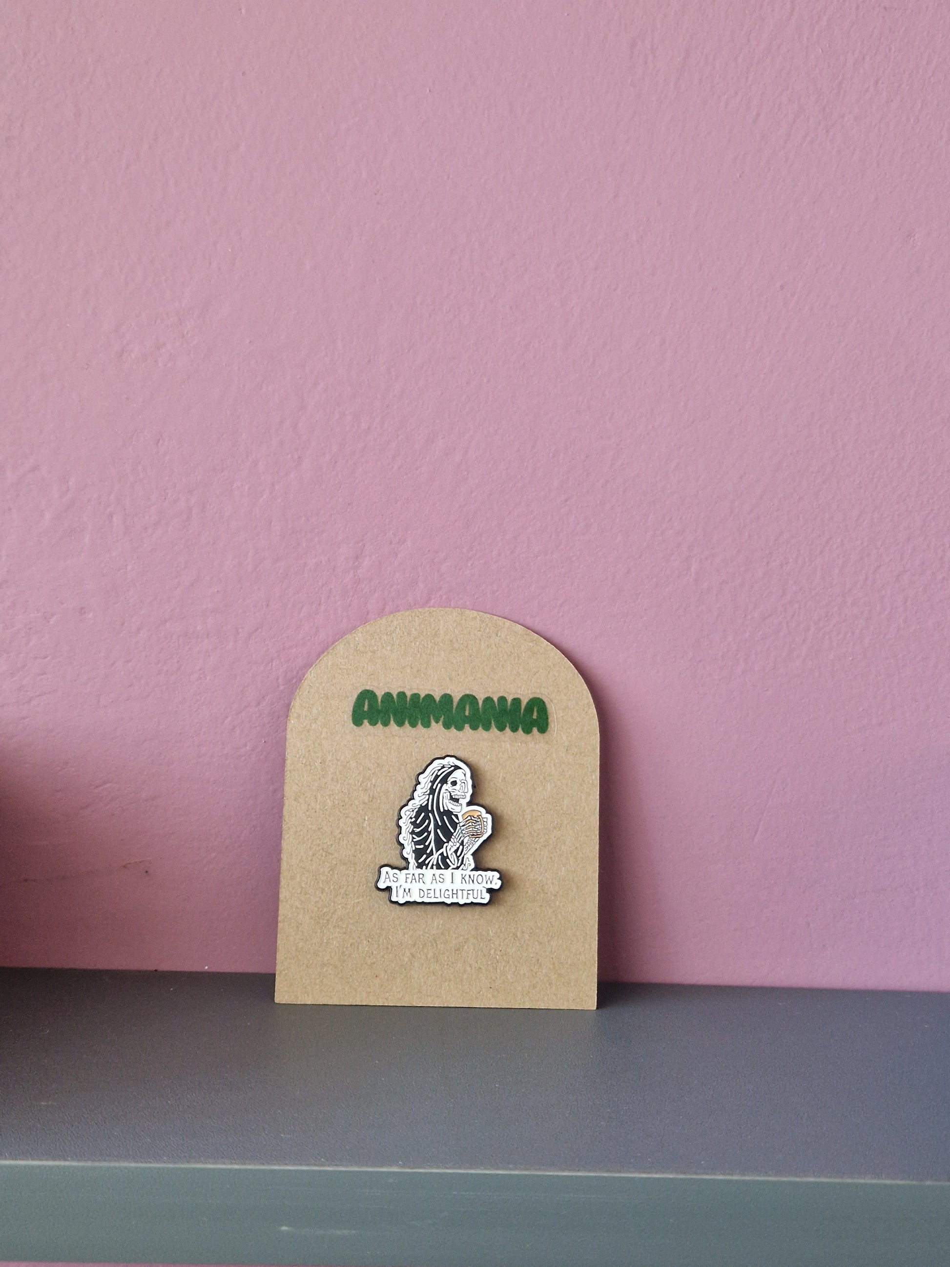 A black and white skeleton holding a cup of coffee with 'as far as i know, i'm delightful' written underneath it. Pin is mounted on a brown cardboard backing with the brand Animania written above it.