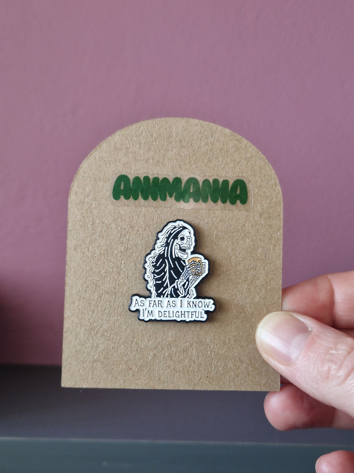 A black and white skeleton holding a cup of coffee with 'as far as i know, i'm delightful' written underneath it. Pin is mounted on a brown cardboard backing with the brand Animania written above it.