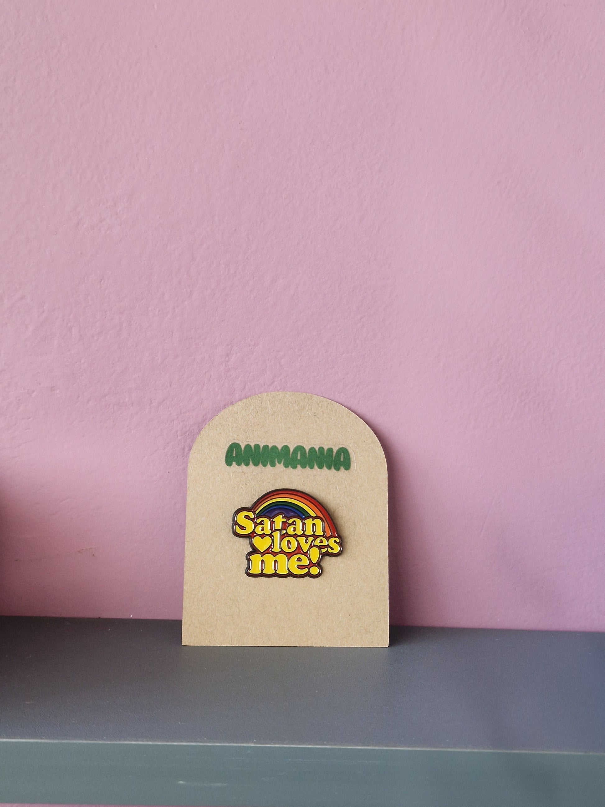 Brightly coloured rainbow pin with 'satan loves me' written underneath it. Pin is mounted on a brown cardboard backing with the brand Animania written above it.