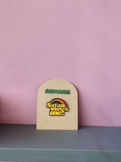 Brightly coloured rainbow pin with 'satan loves me' written underneath it. Pin is mounted on a brown cardboard backing with the brand Animania written above it.
