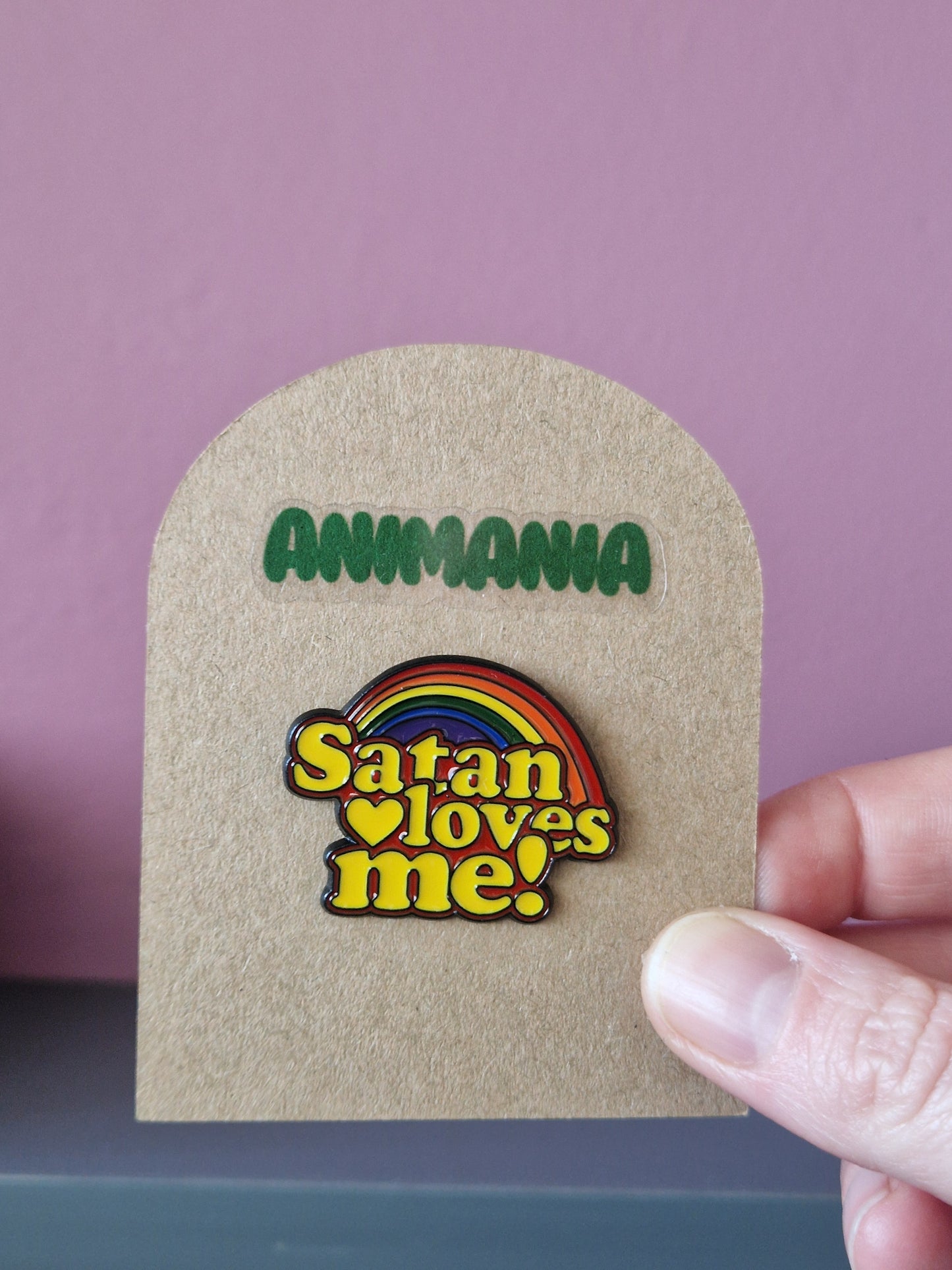 Brightly coloured rainbow pin with 'satan loves me' written underneath it. Pin is mounted on a brown cardboard backing with the brand Animania written above it.