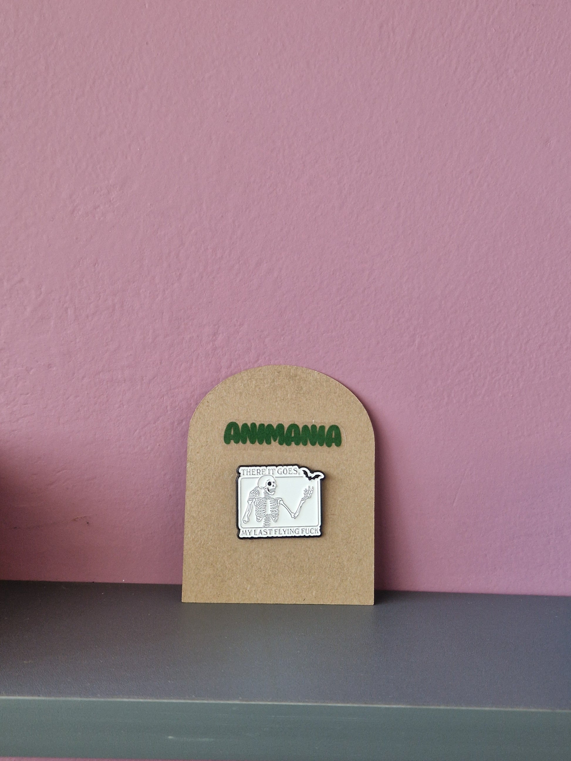 A black and white skeleton & bat pin with 'there it goes, my last flying fuck' written above & underneath it. Pin is mounted on a brown cardboard backing with the brand Animania written above it.
