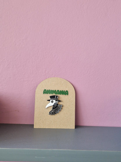 A black and white plague doctor pin. Pin is mounted on a brown cardboard backing with the brand Animania written above it.