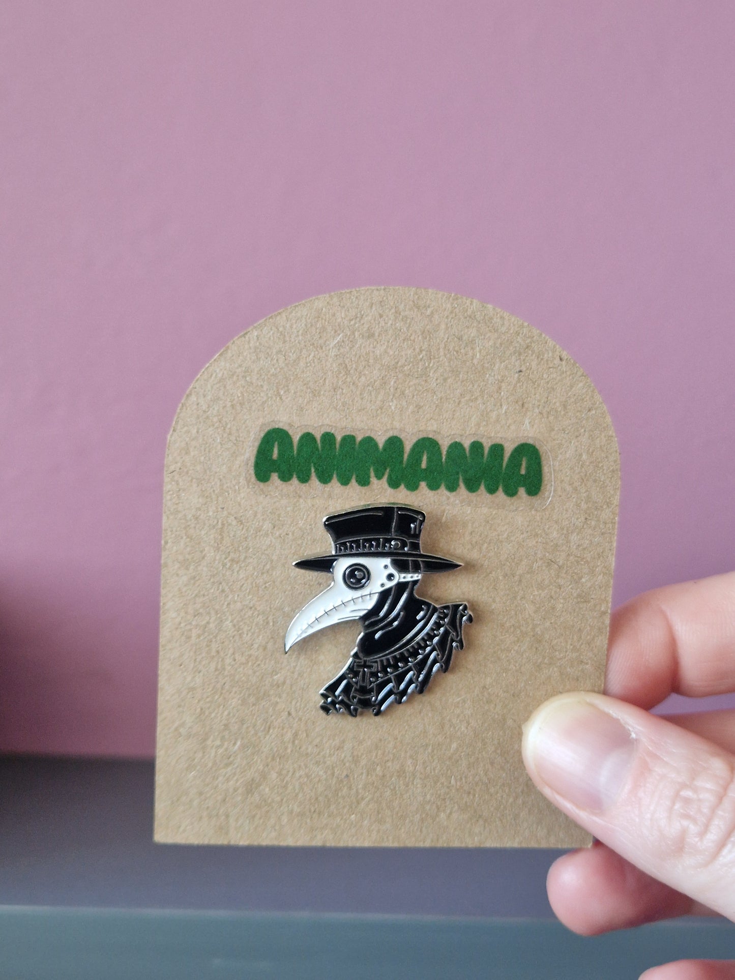 A black and white plague doctor pin. Pin is mounted on a brown cardboard backing with the brand Animania written above it.
